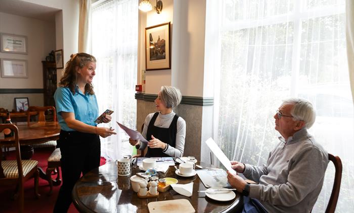 In this image Tracey is wearing a blue shirt with the restaurant windows letting all the light in onto 2 guests sitting at the table ordering their br